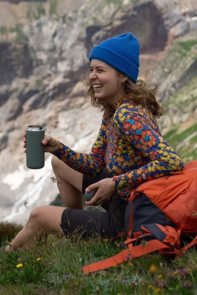 Žena v modré čepici a barevném svetru ze 100% merina vlny sedí na trávě u hory, usmívá se a drží nápoj, vedle má oranžový batoh.