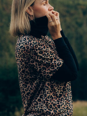 Osoba s krátkými blond vlasy v leopardí košili přes černý rolák z wełny merino 100% stojí venku, oči zavřené, rolák přes ústa.