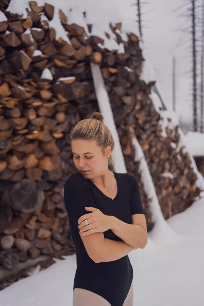 Žena v dámském těle Merino SKIN - černé (135 g) z wełny merino 100% stojí se zkříženýma rukama a zavřenýma očima před hromadou dřeva.